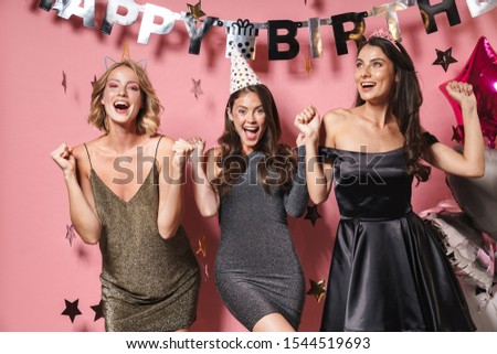 Image of three excited party girls in festive dresses smiling and celebrating birthday isolated over pink background