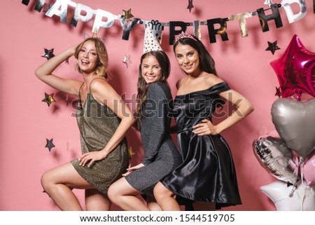 Image of three elegant party girls in festive dresses smiling and celebrating birthday isolated over pink background