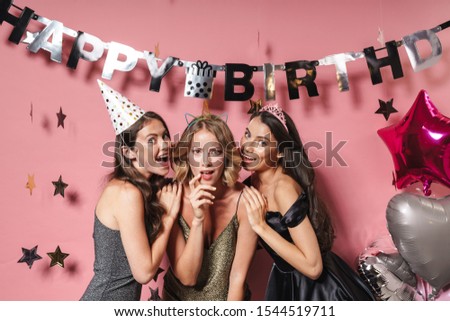 Image of three delighted party girls in festive outfits smiling and celebrating birthday isolated over pink background