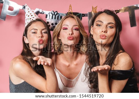 Image of three fashionable party girls in festive outfits blowing air kiss and celebrating birthday isolated over pink background