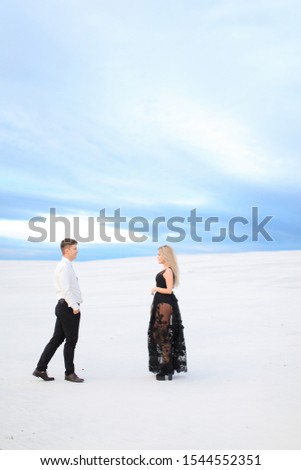 Young man in white shirt and woman wearing black transparent dress walking on snow. Concept of winter photo session, relationship and love, happy couple.