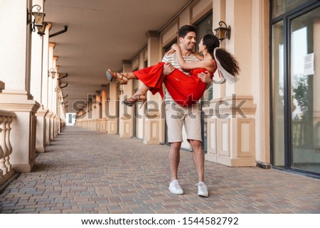 Image of young unshaven man carrying happy woman in hands and smiling while walking near beautiful building