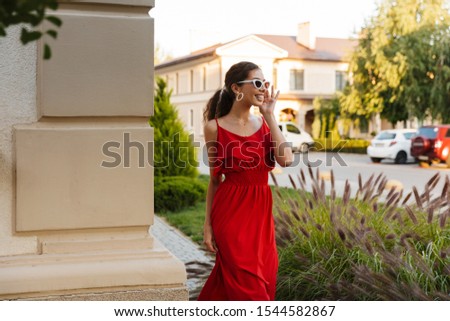 Image of joyful young woman wearing red dress and sunglasses smiling while walking near building outdoors