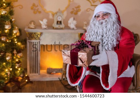 Santa Claus offer wrapped gift box at home by the Christmas tree and fireplace. Merry Holidays and many gifts. Copy space