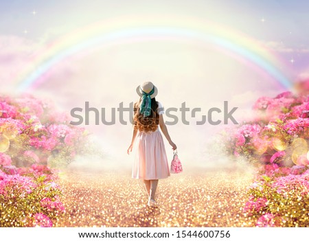 Young lady woman in romantic pink dress, retro hat, bag walking along rose garden path leading to fabulous rainbow unicorn house, flecks of sunlight on road. Tranquil fantasy scene, fairytale hills. Royalty-Free Stock Photo #1544600756