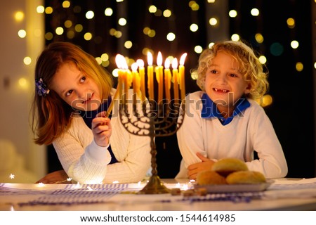 Kids celebrating Hanukkah. Jewish festival of lights. Children lighting candles on a traditional menorah. Boy in a kippah with a dreidel and Sufganiyah doughnut. Israel holiday.