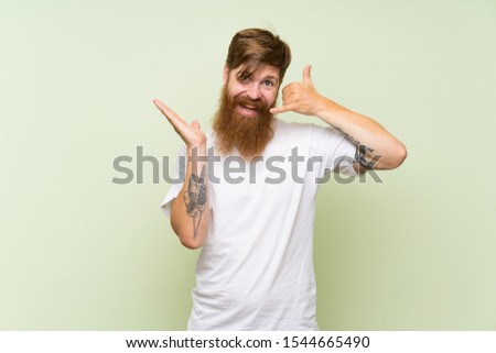 Redhead man with long beard over isolated green background making phone gesture and doubting