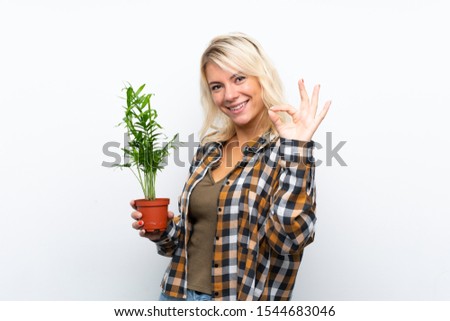 Young blonde gardener woman holding a plant over isolated white background showing ok sign with fingers