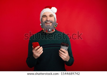 Happy bearded man wearing santa claus hat holding smartphone and cup of coffee to go