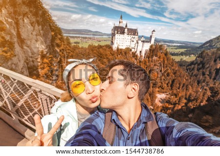 A happy Asian girl and a European traveler embrace and kiss during their honeymoon trip in the background of the famous Neuschwanstein castle.