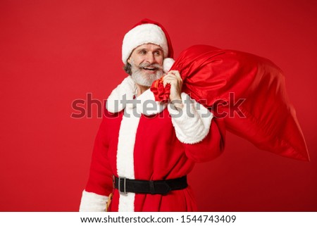 Cheerful elderly gray-haired mustache bearded Santa man in Christmas hat posing isolated on red background. Happy New Year 2020 celebration holiday concept. Mock up copy space. Hold bag with presents