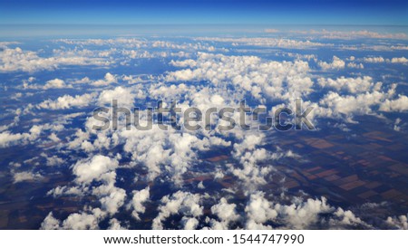 Flying over beautiful heaps of white clouds moving softly from right to left on the bright blue sky, and countryside landscapes far beneath. Picturesque view from the airplane window.