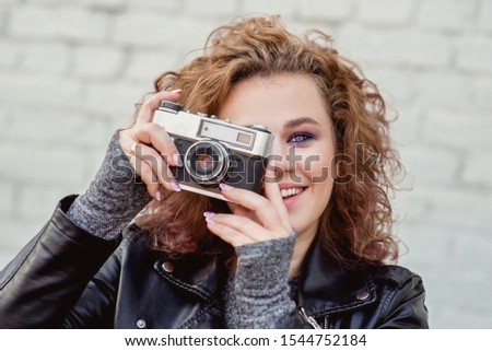 portrait of young beautiful cheerful woman with red curly hair taking pictures on film oldfashioned camera on white bricks wall background. Film stylization