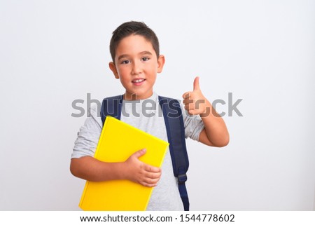 Beautiful student kid boy wearing backpack holding book over isolated white background happy with big smile doing ok sign, thumb up with fingers, excellent sign