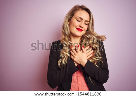 Young beautiful business woman wearing elegant jacket standing over pink isolated background smiling with hands on chest with closed eyes and grateful gesture on face. Health concept.