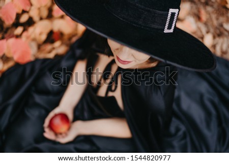 Unrecognizable girl as witch in black hat holding red apple as symbol of temptation, poison. Fairy tale concept, halloween, cosplay.