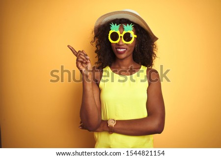 African american woman wearing funny pineapple sunglasses over isolated yellow background with a big smile on face, pointing with hand and finger to the side looking at the camera.