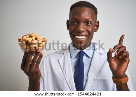 African american doctor man holding bowl with peanuts over isolated white background very happy pointing with hand and finger to the side