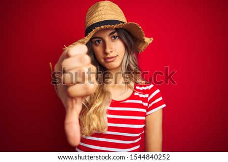 Young beautiful woman wearing summer hat over red isolated background with angry face, negative sign showing dislike with thumbs down, rejection concept