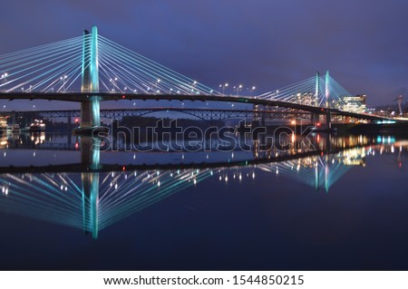 The Tillikum Bridge crosses the Willamette River in Portland, Oregon. The lights from the city's modern structures are reflected in the calm waters. 