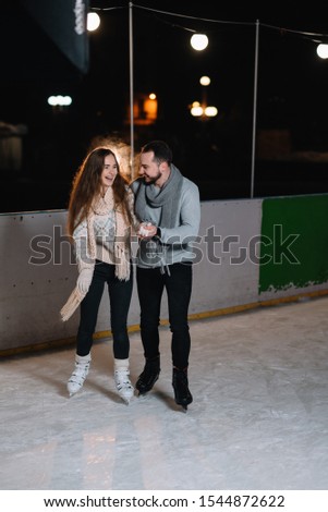 Man and woman young family Happy smiles hold hands skate on winter rink at night, with bokeh lights.