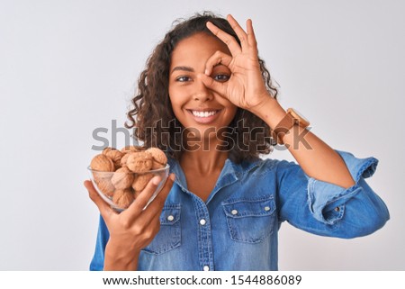 Young brazilian woman holding bowl with walnuts standing over isolated white background with happy face smiling doing ok sign with hand on eye looking through fingers