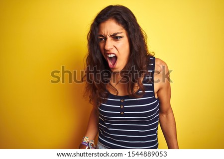 Young beautiful woman wearing striped t-shirt standing over isolated yellow background angry and mad screaming frustrated and furious, shouting with anger. Rage and aggressive concept.