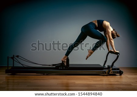 Young girl doing pilates exercises with a reformer bed. Beautiful slim fitness trainer on a reformer gray background, low key, art light, copy space advertising banner Royalty-Free Stock Photo #1544894840