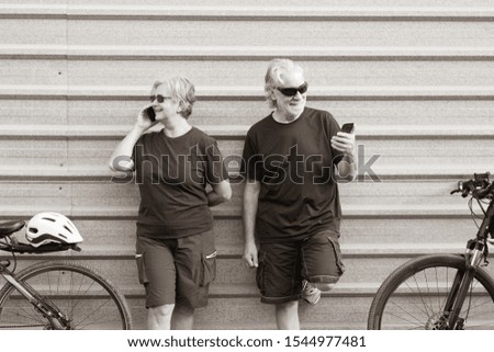 Elderly couple of people looks at the cellphone outdoor against a gray metal panel. Resting after activity with electric bicycle. Healthy lifestyle. Yellow helmet, black clothes.