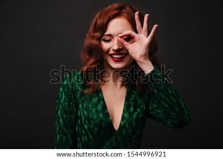 Shy but joyful girl enjoys great Christmas mood and poses funny. Photo on black background of young lady with red lipstick showing sign ok