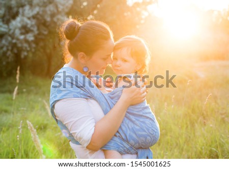  Touching mother and baby girl portrait in sunset light using child wrap, sling, carrier, canvas blue outdoors, baby wearing concept Royalty-Free Stock Photo #1545001625