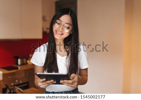 Positive Asian girl in white T-shirt and fashionable glasses posing with computer tablet on background of kitchen
