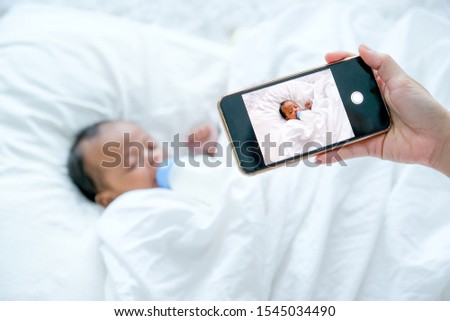 Mother hand hold mobile phone to take a photo of newborn baby during she is sleeping on white bed.