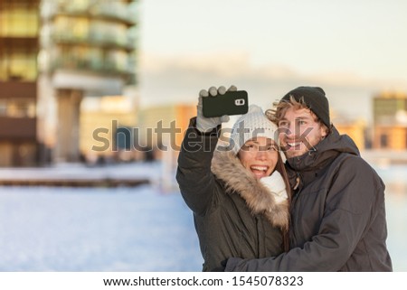 Selfie couple happy winter travel walk tourists taking photo with phone on city street panorama lifestyle. Asian woman, Caucasian man wearing knit hats and jackets.