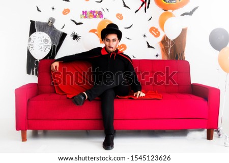 Handsome guy is wearing a costume and dressed in Dracula's face,relaxed, sitting on the red sofa in the living room, decorated with balloons and stickers at the annual Halloween festival.