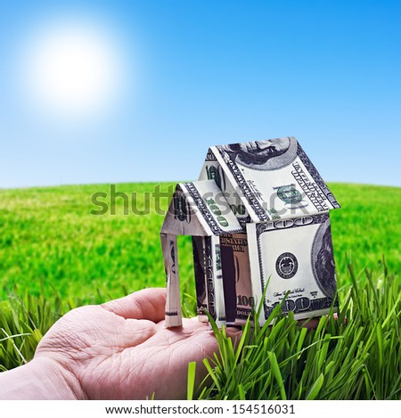 House made of money in hand on background of green field