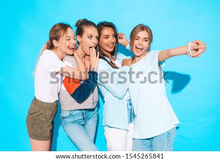 Four young smiling hipster women in summer clothes.Girls taking selfie self portrait photos on smartphone.Models posing near blue wall in studio,Female showing positive face emotions