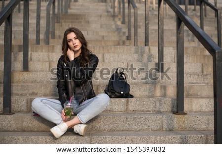 Portrait of european young beautiful smiling woman with dark straight hair sitting on stairs in black leather jacket and blue jeans with drink in hand
