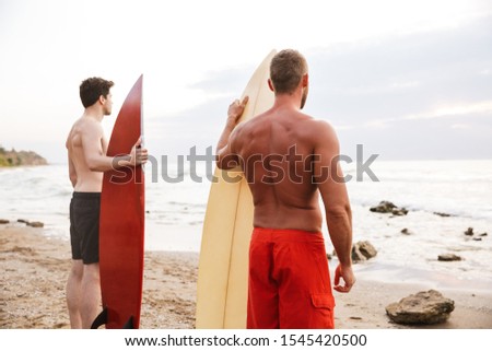 Back view picture of a young two men surfers friends with surfings on a beach outside.