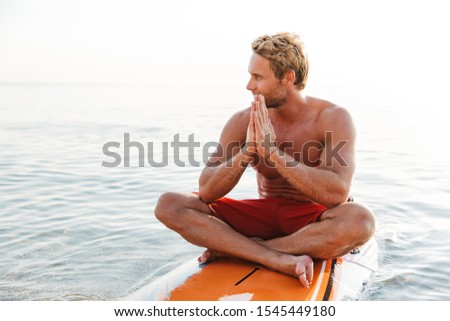 Image of a concentrated handsome young man surfer with surfing desk at the beach sea meditate.