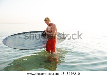 Picture of attractive concentrated young man surfer with surfing desk at the beach sea.