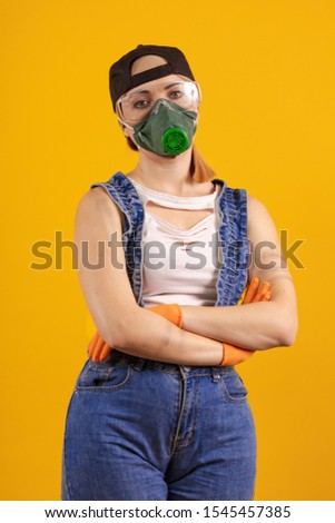 Builder girl in respiratory mask in denim overalls and a black cap posing on a yellow background