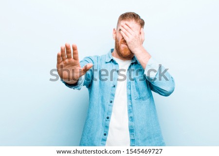 young red head man covering face with hand and putting other hand up front to stop camera, refusing photos or pictures against soft blue background