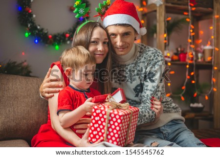 Mom and dad give their son a gift in a red box. A young family with a child celebrates Christmas and New year in a cozy home environment
