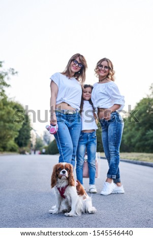 Three young girls, wearing blue jeans and white t-shirts, walking with small dog in city park in summer. Family fun leisure time with cavalier king charles spaniel. Pretty women posing for picture.