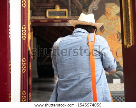 Tourist man wearing hat, walking and photographing wall painting temples area in Bangkok. He uses the orange shoulder bag. Standing back to take pictures. Enjoy and fun visiting temples in Thailand.