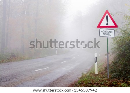 Street sign "Achtung Nebel" ahead a foggy road in autumn meaning "Warning fog" 