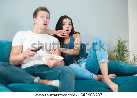 young couple beautiful brunette woman and handsome man watching scary movie on the sofa eating pop corn looking frightened cozy lifestyle