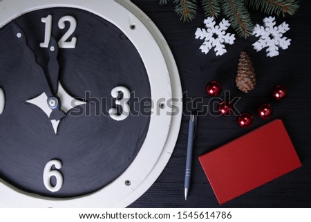 A black and white wooden watch, a smile made of twigs and fir cone, penoplast flakes, new year's toys, a pen and a red book all arranged on a blackboard. Top view. Place for advertisement.
