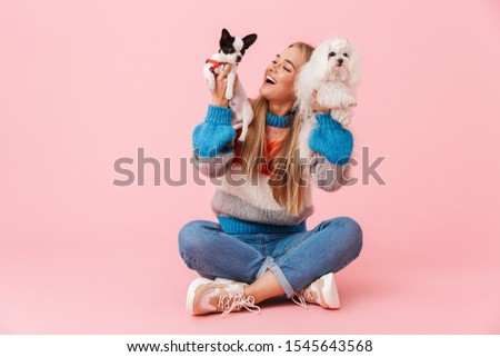 Cute lovely girl wearing sweater sitting with legs crossed with her pet chihuahua and lapdog isolated over pink background